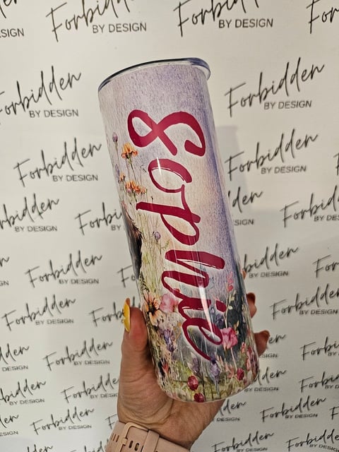 Hand holding a customized tumbler with pink flowing script design and floral elements against a Forbidden By Design branded backdrop
