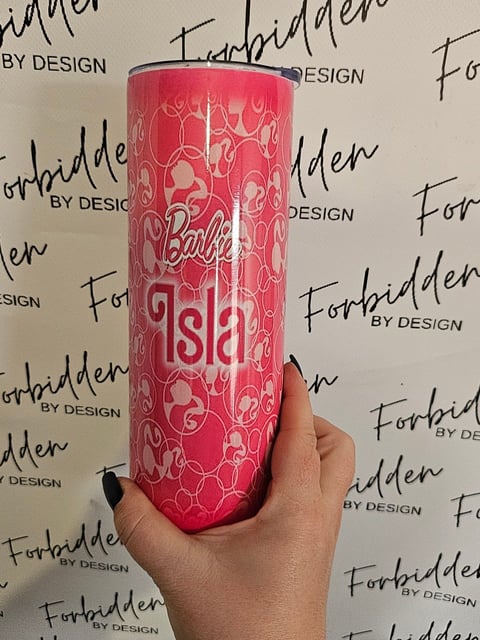 Hand holding a pink Barbie-themed tumbler with decorative pattern against a Forbidden by Design backdrop