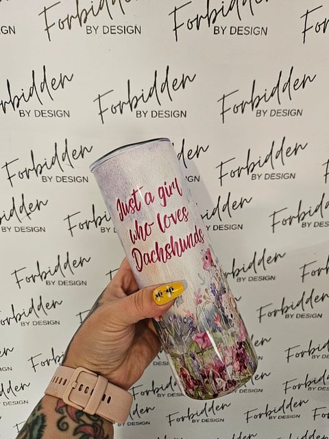 Hand holding a white tumbler with red text reading Just a girl who loves Dachshunds against a Forbidden By Design branded background