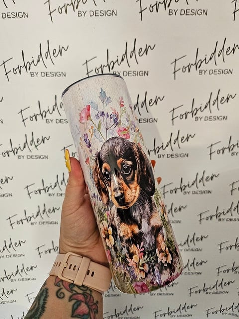 Hand holding a white cylindrical mug decorated with a portrait of a black and tan dog surrounded by colorful flowers