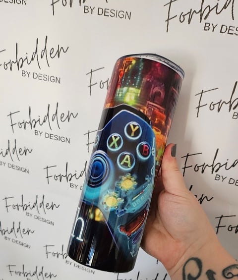 Hand holding a custom tumbler with Xbox controller and colorful gaming graphics against a Forbidden by Design branded backdrop